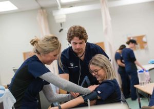 Two students wearing scrubs apply blood pressure cuff to third student