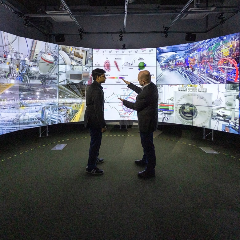Bhavesh and Robert are shadowed by a giant rounded TV screen, which has a number of scientific-looking photos projected onto the screen.