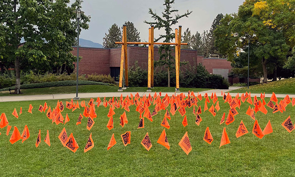 A collection of orange flags with hummingbirds printed on them in front of the Story Poles.