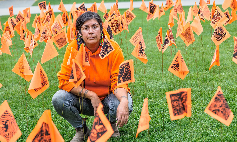 Tania Willard wears an orange sweater and kneels amongst dozens of orange flags inserted into the ground.