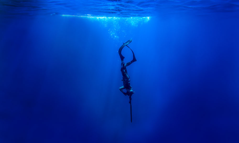 An underwater photo of a diver diving down into the depths of the ocean holding a spear-like object straight down. Bubbles and light rays of blue are seen at the top of the photo, signifying the surface.