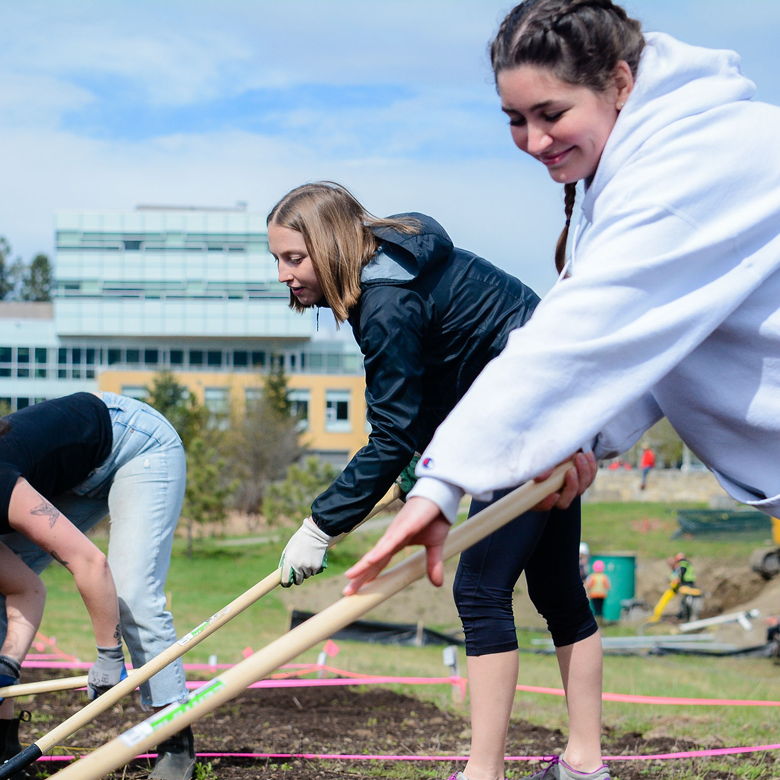 Three students are pictured raking the dirt and smiling, while the EME building on UBC Okanagan's campus stands in the background