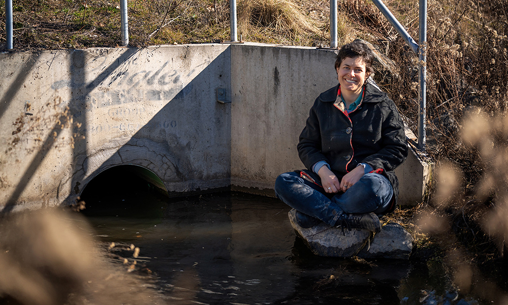 Researcher madeline Donald sits on a rock nestled in a creek culvert. The space looks urban and industrial but the sun shines on the creek, making it glisten
