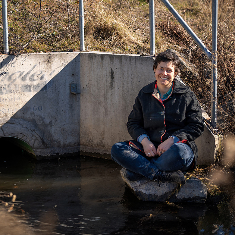 Researcher madeline Donald sits on a rock nestled in a creek culvert. The space looks urban and industrial but the sun shines on the creek, making it glisten