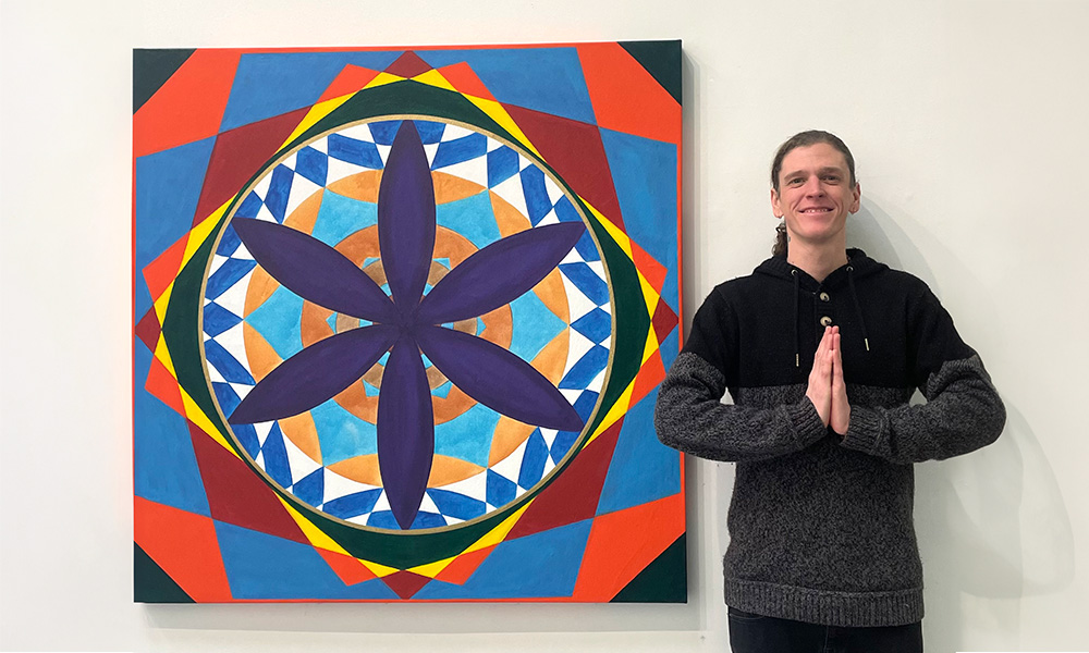 Undergraduate student Asana Hughes holds his hands in front of his chest in a prayer while he stands next to one of his paintings of a mandala.